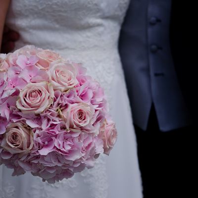 Nahaufnahme eines rosa Brautstraußes vor einem festlich gekleideten Brautpaar